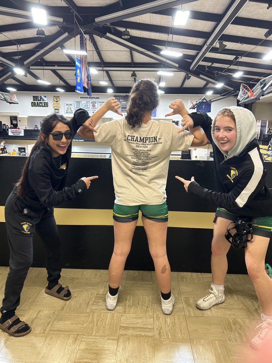 Ryleigh Rangel and Khloe Kiefer point excitedly at Isabel Sayre's champion shirt. 